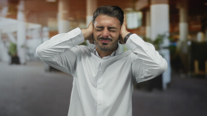 Man in white shirt holds hands over ears in building foyer under natural light as he winces from...