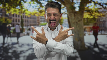 Young hispanic man crossing hands into rock horns gesture with open mouth energetically yelling in...
