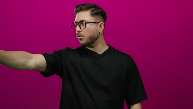 Young man with beard and glasses in black shirt points confidently against vivid pink background conveying focus and determination