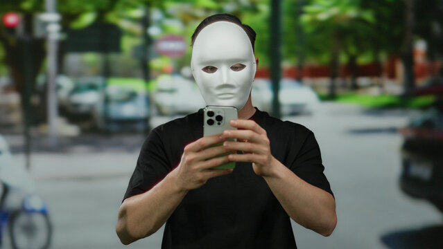 Masked man holding smartphone on busy city street surrounded by blurred traffic and greenery showcasing urban anonymity and modern society.