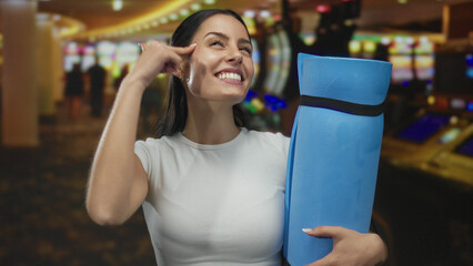 Woman pointing at head with finger while holding blue mat in busy casino with blurred slot machines...