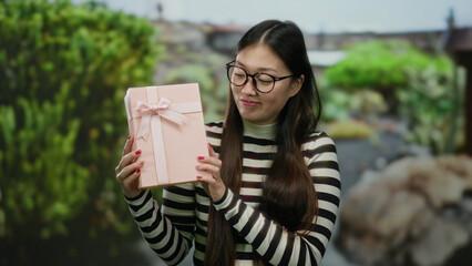Woman holding pink gift outdoors in a park setting, wearing glasses and striped sweater with a...