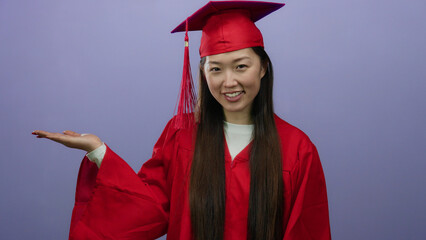 Graduate woman in red cap and gown against a purple background with hand raised for display,...