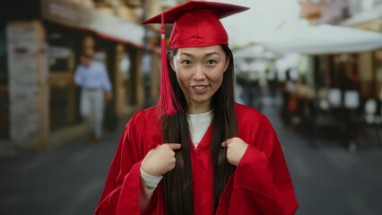 Woman in red graduation gown and cap stands on urban street pointing at herself with a blurred city...