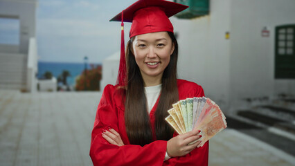 Woman in red graduation robe holds philippine pesos on an urban street, symbolizing achievement and...