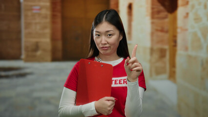 Woman holding clipboard in outdoor university setting pointing with expressive gesture while volunteering in casual red shirt against old college building wall.