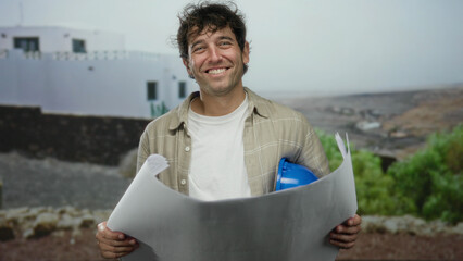 Hispanic man holding blueprints and wearing a hardhat stands outdoors on a street with a serene...