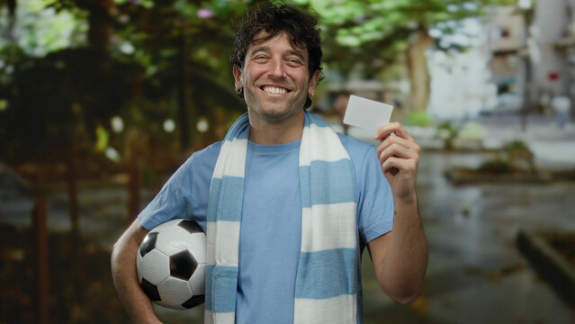 Hispanic man holding soccer ball and card outside city street wearing scarf in urban setting.