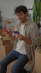 Hispanic man smiling while using smartphone and credit card in new home surrounded by boxes...