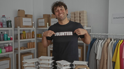 Hispanic man smiling proudly in a volunteer shirt stands in a donation room with shelves of...