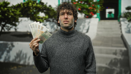 Hispanic man holding czech koruna banknotes outdoors on a city street appears thoughtful,...