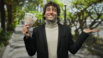 Hispanic man smiling outdoors holding argentine pesos in a city setting with his hand gesturing to...