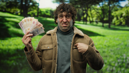 Hispanic man outdoors in a park expresses incredulity while holding turkish lira banknotes,...