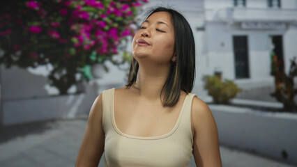 Young chinese woman breathing deeply outdoors in a city street surrounded by vibrant flowers and white buildings, captured in a serene moment of mindfulness and tranquility.