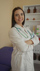 Young hispanic woman doctor with stethoscope smiles confidently in a clinic room with medical...