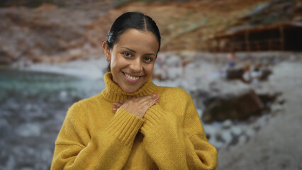 Woman smiling warmly with closed eyes and hands on chest, wearing a yellow sweater by the seaside...