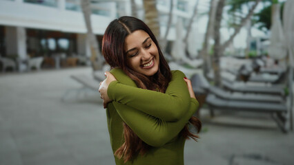 Woman smiling outdoors embracing herself on a sunny day on a busy street with trees and people in the background depicting warmth and happiness
