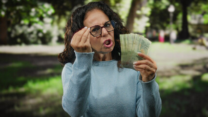 Middle-aged woman in park holding argentine pesos, wearing glasses and sweater, expressing anger...