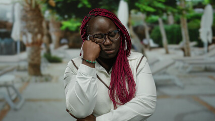 Fototapeta premium Woman standing outdoors in a city street with her hand on chin looking thoughtful, wearing glasses and braids, surrounded by trees and urban scenery.