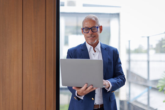 Happy middle aged business man ceo wearing suit standing in office using laptop. Smiling mature businessman professional executive manager looking away thinking working on tech device.