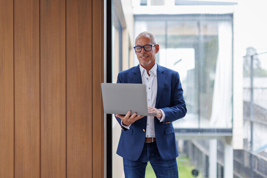 Happy middle aged business man ceo wearing suit standing in office using laptop. Smiling mature businessman professional executive manager looking away thinking working on tech device.