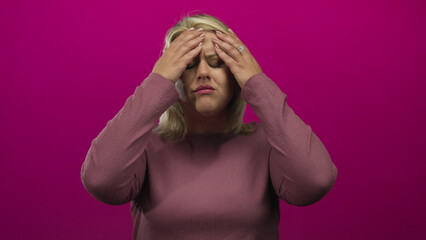 Young blonde woman in mauve sweater presses temples with furrowed brow and closes eyes in studio;...