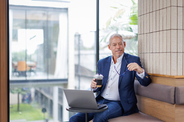 Handsome middle aged in casual clothes and stylish eyeglasses holding paper cup with takeaway drink and working on laptop in modern office environment, successful business concept
