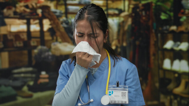 Woman doctor in uniform with stethoscope sneezing indoors at a retail clothing store setting. - Powered by Adobe