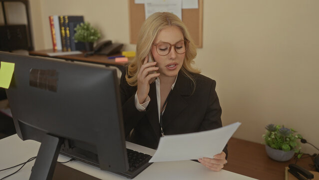 Woman talking on a phone in an office while reviewing documents, showcasing a productive work environment with a computer desk setup indoors and professional attire.