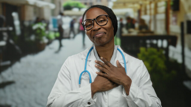 Woman smiling with closed eyes wearing glasses and lab coat, outdoors in a vibrant urban street setting, expressing gratitude with a stethoscope around her neck.
