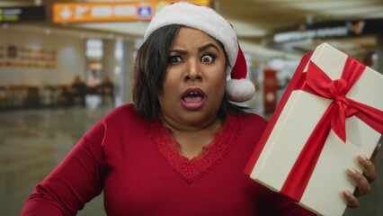 Woman in airport terminal with a christmas gift wearing santa hat and red sweater, showing surprise...