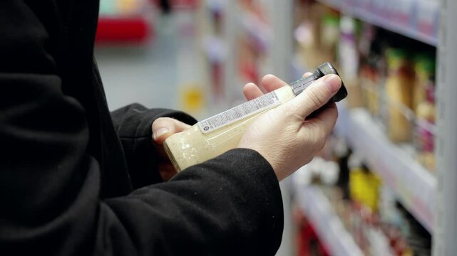 Focused woman reading product label, checking nutritional details while comparing sauce ingredients in supermarket aisle, highlighting health conscious shopping behavior