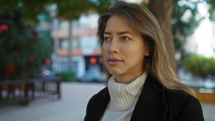 Woman in urban park wearing sweater and jacket looking thoughtful surrounded by trees and buildings...