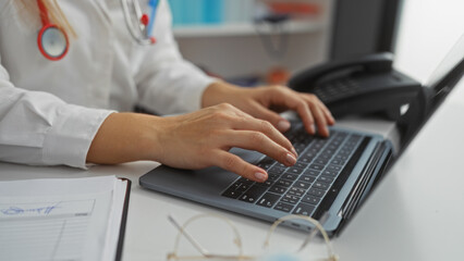 Female doctor typing on a laptop in a hospital office with stethoscope on desk indicating a medical...