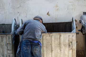 A man bends over two trash bins in an alley, looking for items of value under the warm afternoon...