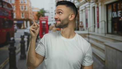 Young hispanic man in urban london street points upward with a smile, surrounded by classic red bus...