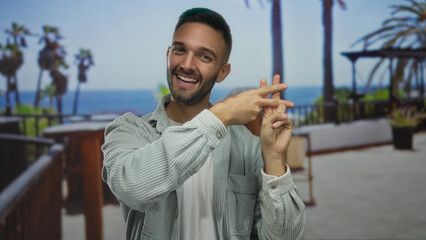 Young hispanic man making hashtag gesture on sunny seaside promenade with ocean view and palm trees, exuding happiness and relaxation outdoors.