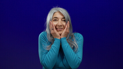 Woman with gray hair wearing blue sweater looks surprised against a vivid blue background,...