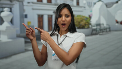 Woman smiling in the outdoor street wearing a name badge, pointing confidently with a friendly...