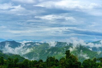 Scenic panoramic view of lush green mountains with floating mist under dramatic cloudy skies, capturing the tranquil beauty of nature after rainfall.