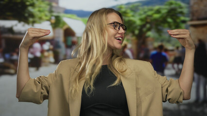 Woman gesturing while talking on a city street, wearing a beige jacket, surrounded by blurred pedestrians in a vibrant outdoor setting.