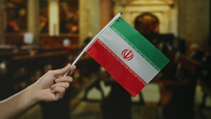 Hand holding iranian flag indoors, with a background of a church setting and people, highlighting cultural connection and diversity.