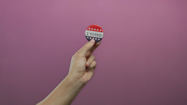 Caucasian man holds a voting badge against an isolated pink background, symbolizing civic engagement and democratic participation in an election.
