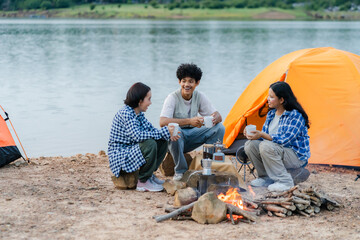 A group of friends sitting by the lake around a campfire, chatting and brewing coffee in front of an orange tent. Warm, relaxed outdoor camping atmosphere close to nature.