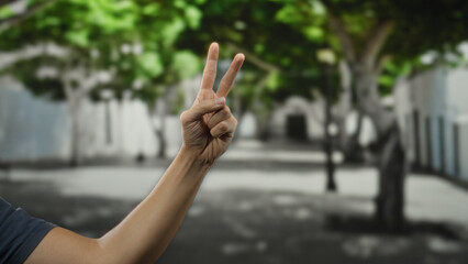 Man hand showing peace gesture in outdoor street setting with blurred city background and green trees.