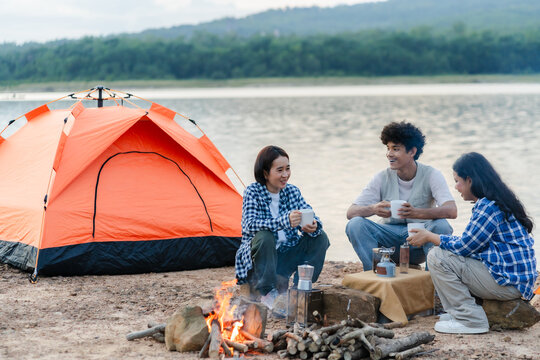 A group of friends sitting by the lake around a campfire, chatting and brewing coffee in front of an orange tent. Warm, relaxed outdoor camping atmosphere close to nature. - Powered by Adobe