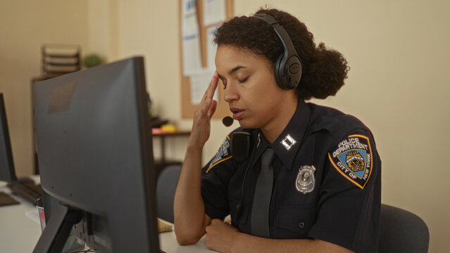 Hispanic woman police officer in uniform communicates at a desk in an indoor station, using a headset and computer for efficient task management.