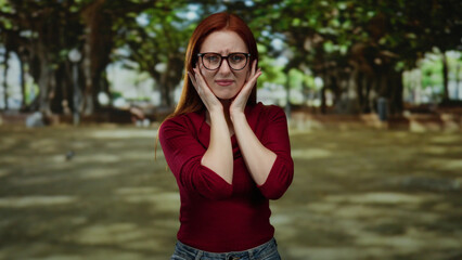 Woman with red hair in a park covering ears from loud noise outdoors, captures a young adult's...
