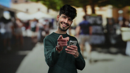 Young man smiling while pointing at camera on urban street holding smartphone illustrates vibrant...