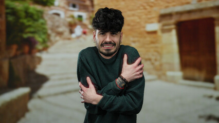 Young man shivering on an urban street, wearing a green sweater, expressing cold in a rustic town...